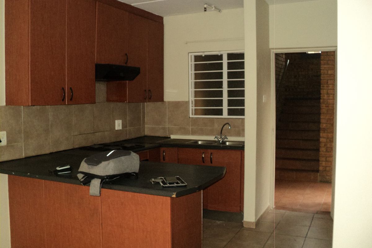 A functional kitchen featuring laminate cabinetry, dark countertops, a tiled backsplash, and a stainless steel sink with a view toward an adjacent stairwell.