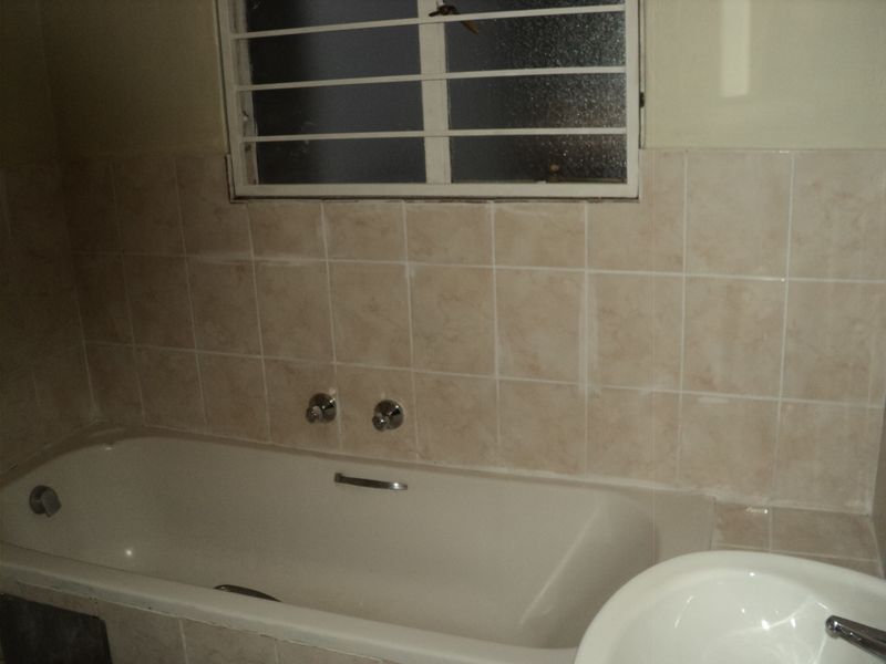 A dated bathroom featuring a built-in bathtub with wall-mounted taps, ceramic wall tiling, and a window with security bars.