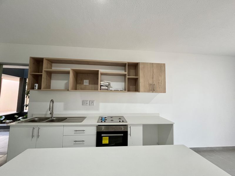 A modern, clean kitchen featuring light wood cabinetry, white stone countertops, a built-in oven, gas hob, and a stainless steel sink.