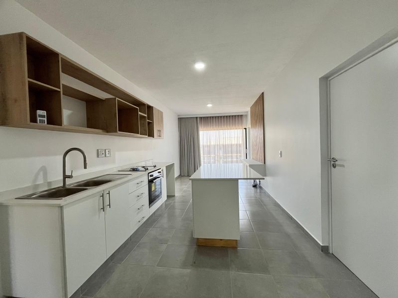 Modern open-plan kitchen featuring white cabinetry, a central island, built-in oven, and sleek tiled flooring, leading to a bright living area.
