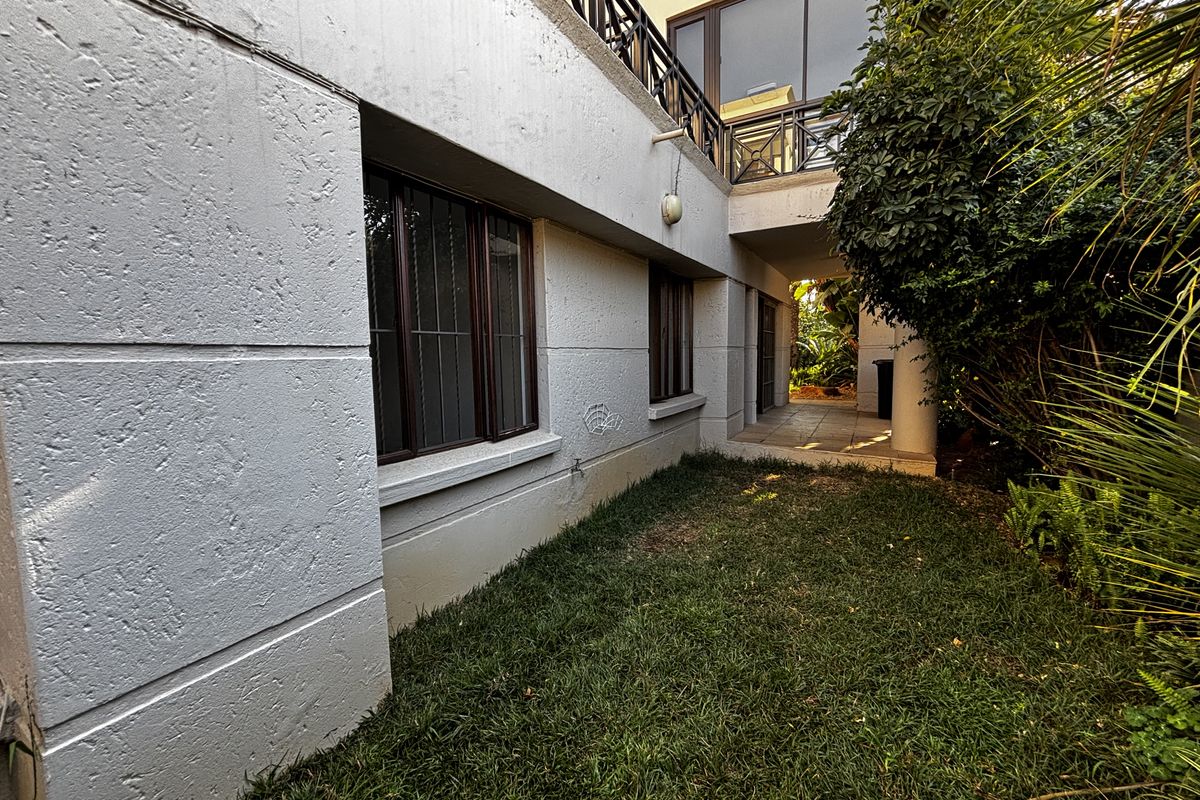 Rear exterior view of the apartment featuring a small lawn area, tiled patio space, and security bars on the windows.