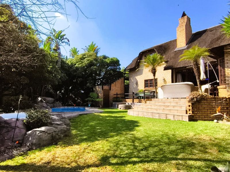 Rear view of a charming thatched-roof house featuring a brick facade, elevated patio steps, and a landscaped garden with a swimming pool.
