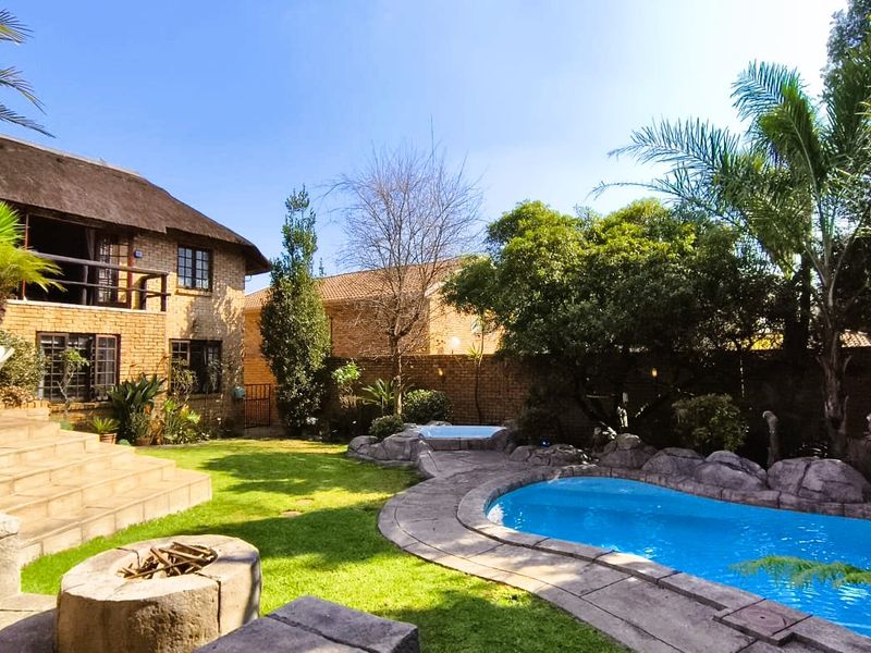 A well-maintained outdoor area featuring a swimming pool with rock surrounds, a stone fire pit, and a multi-story brick home with a thatched roof.
