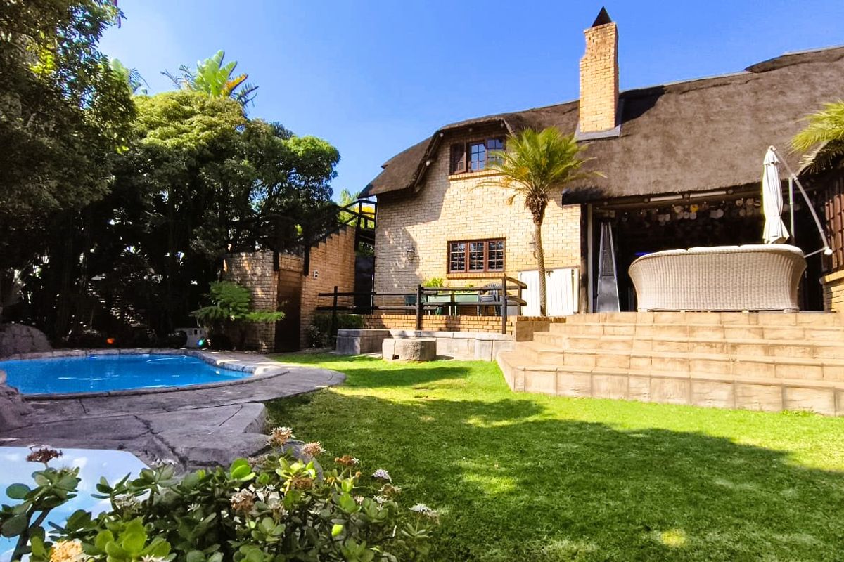 A charming rear exterior view featuring a thatched-roof house, a well-maintained garden, and a private swimming pool with stone surround.