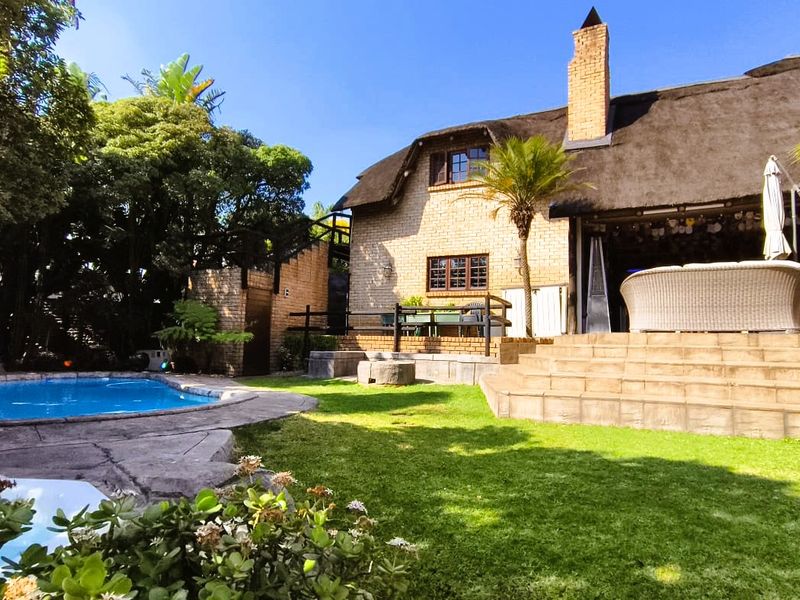 A charming rear exterior view featuring a thatched-roof house, a well-maintained garden, and a private swimming pool with stone surround.