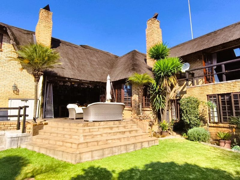 Rear view of a traditional brick house featuring a thatched roof, raised patio area, and an upper-level timber balcony overlooking the garden.