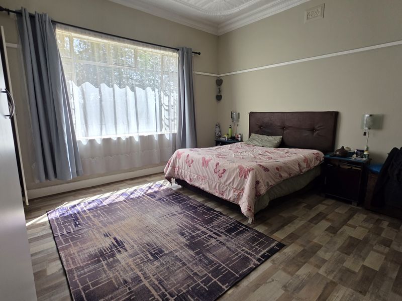 A spacious bedroom featuring laminate flooring, a large window with natural light, and classic architectural details including a ceiling rose and picture rail.