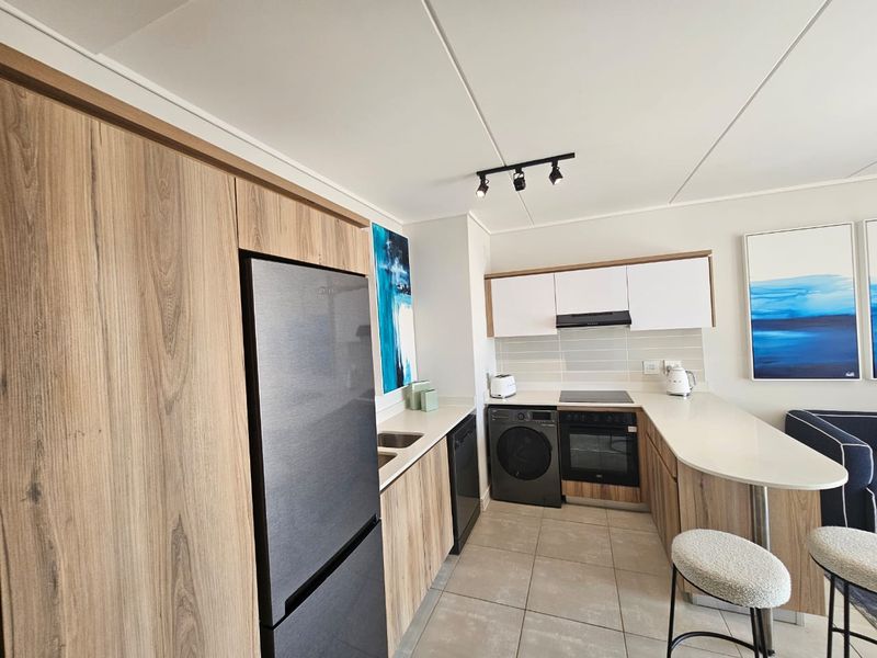 Modern kitchen featuring wood-grain cabinetry, stone countertops, integrated oven, electric hob, and a breakfast bar, finished with neutral tiled flooring.