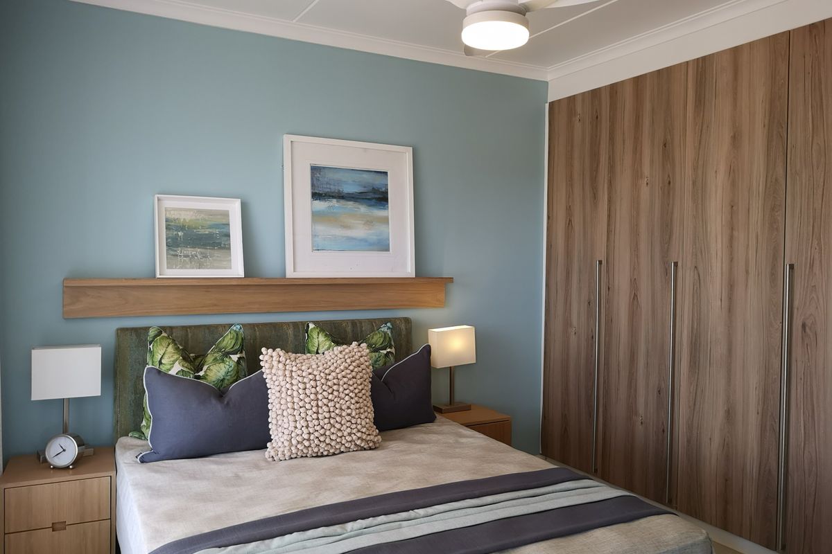 A well-maintained bedroom featuring built-in wood-finish cupboards, a modern ceiling fan, and a decorative floating shelf against a blue accent wall.