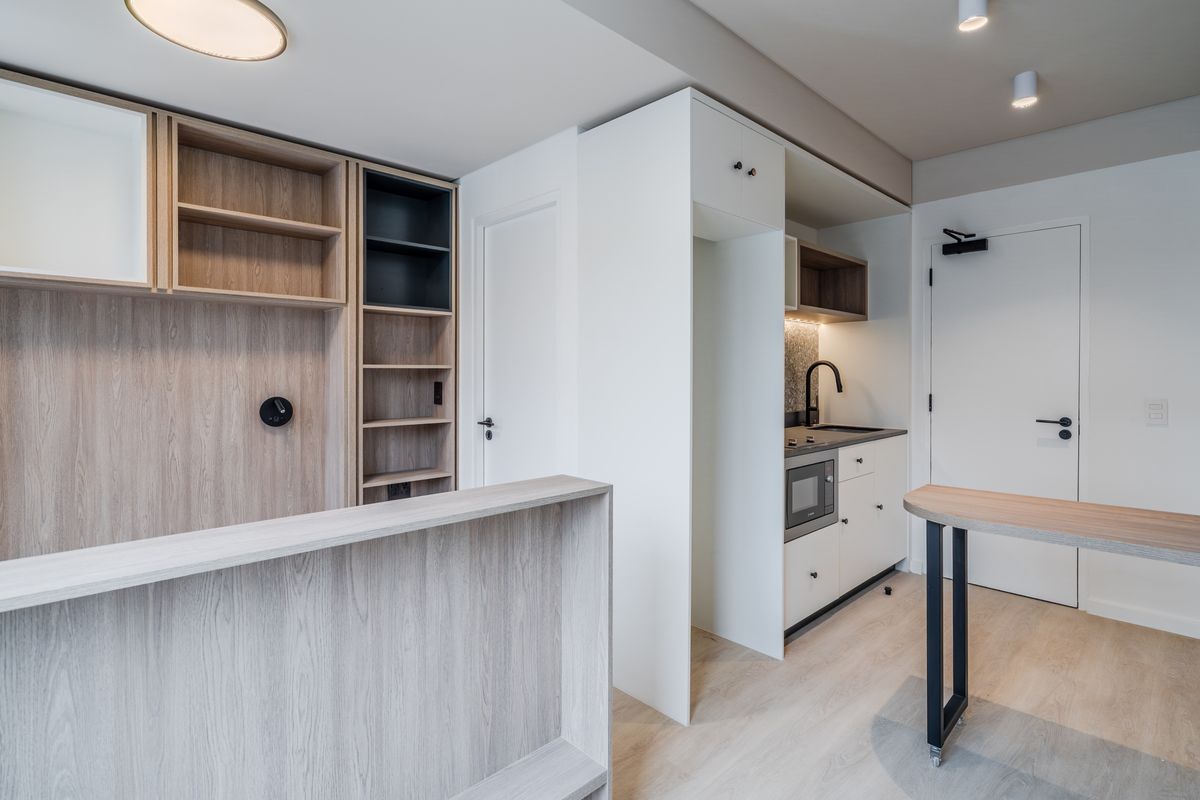 Modern open-plan living area featuring light wood-tone cabinetry, a compact kitchenette with integrated appliances, and neutral finishes throughout.