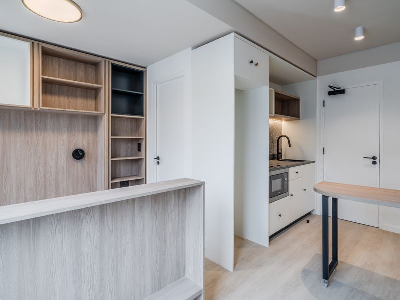 Modern open-plan living area featuring light wood-tone cabinetry, a compact kitchenette with integrated appliances, and neutral finishes throughout.