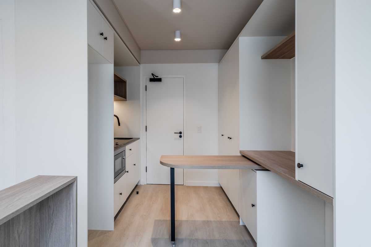 A modern, compact kitchen featuring clean white cabinetry, integrated appliances, wood-tone countertops, and a functional built-in workspace with recessed ceiling lighting.