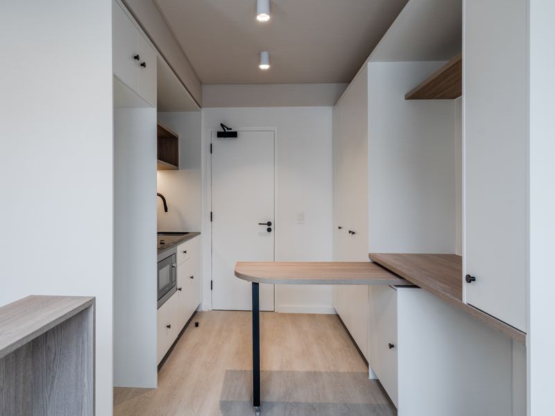 A modern, compact kitchen featuring clean white cabinetry, integrated appliances, wood-tone countertops, and a functional built-in workspace with recessed ceiling lighting.