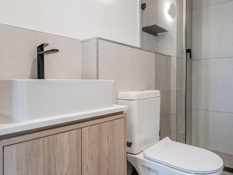 Modern bathroom featuring a vessel sink with black faucet, vanity cabinet, toilet, and a glass-enclosed shower with neutral wall tiling.