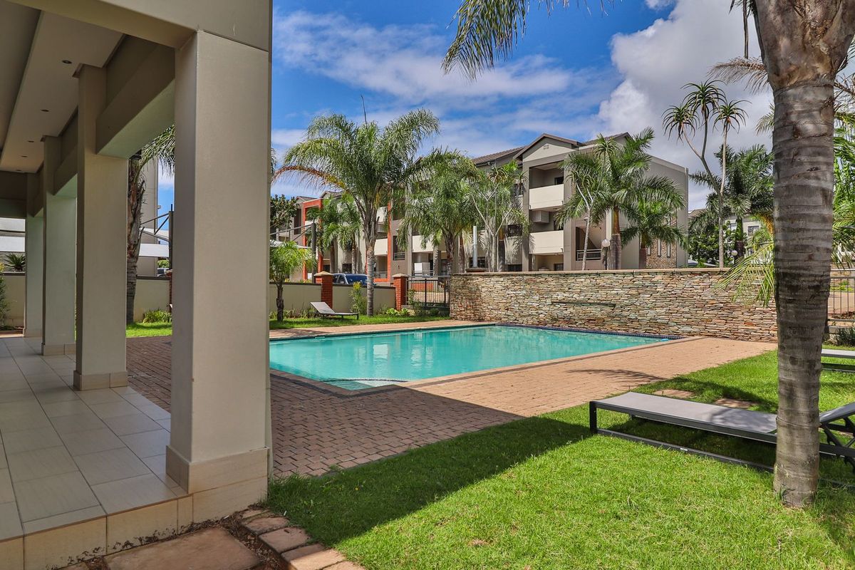 A well-maintained communal swimming pool area featuring a stone feature wall, paved decking, and a covered patio space within an apartment complex.