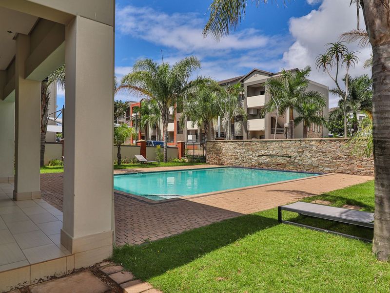 A well-maintained communal swimming pool area featuring a stone feature wall, paved decking, and a covered patio space within an apartment complex.
