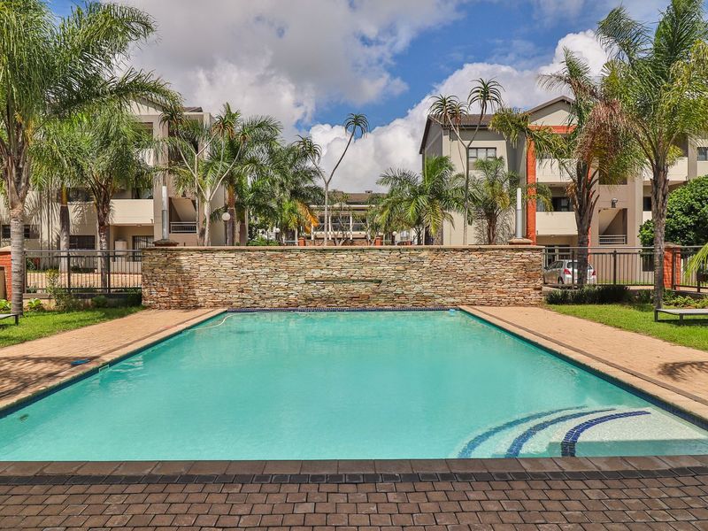 A well-maintained communal swimming pool area featuring a decorative stone wall, paved decking, and lush surrounding landscaping within an apartment complex.