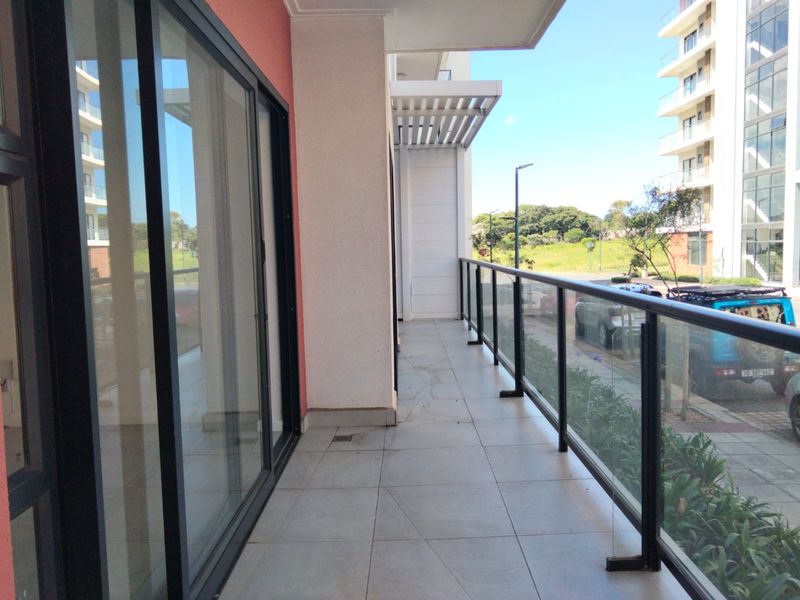 A spacious apartment balcony featuring a modern glass balustrade, tiled flooring, and sliding door access, offering an open outdoor area.