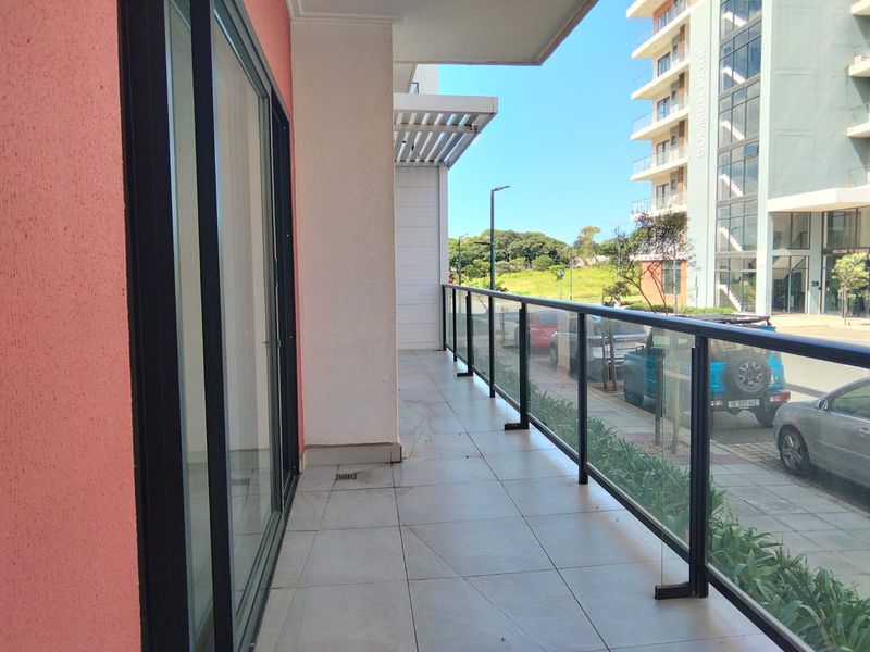 A modern, tiled balcony featuring a sleek glass balustrade and overhead pergola, offering an open outdoor space accessible via large sliding glass doors.