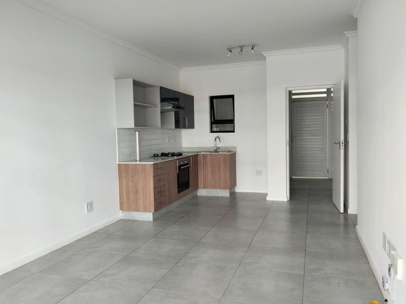 An open-plan kitchen area featuring modern wood-finish cabinetry, a gas hob, built-in oven, and neutral tiled flooring in a clean, vacant apartment space.