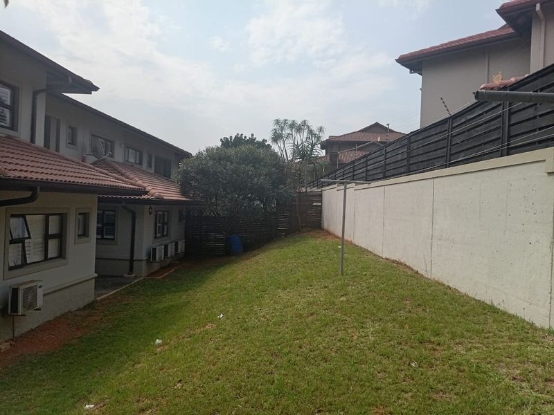 A rear garden area featuring a sloped lawn, boundary wall with electric fencing, and exterior wall-mounted air conditioning units.