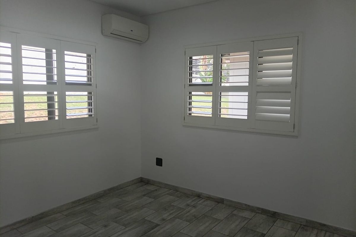 A bright, empty bedroom featuring modern tiled flooring, plantation shutters on the windows, and a wall-mounted split-system air conditioning unit.