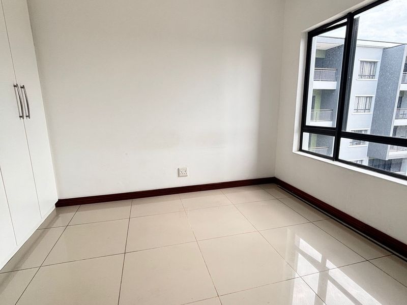 A bright, empty bedroom featuring neutral tiled flooring, built-in cupboards, and large windows providing natural light.