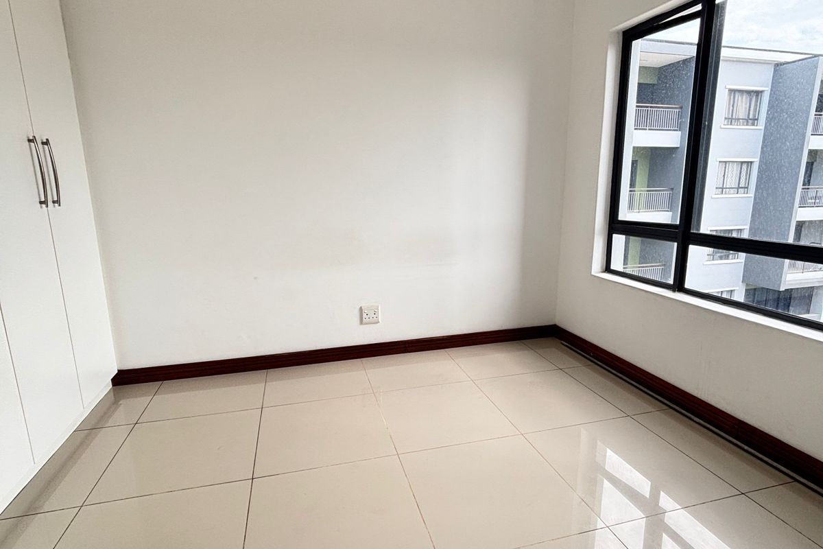 A bright, empty bedroom featuring neutral tiled flooring, built-in cupboards, and large windows providing natural light.