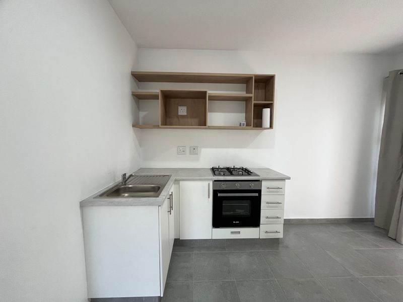 A clean, modern kitchen featuring white cabinetry, light wood open shelving, a built-in oven with gas hob, and grey tiled flooring.