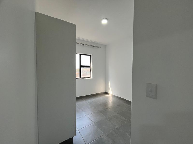 A bright, empty bedroom featuring neutral grey floor tiles, clean white walls, and an aluminium-framed window with a fitted curtain rail.