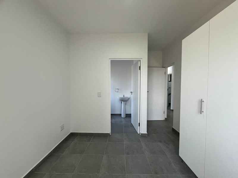 A clean, empty bedroom featuring neutral tiled flooring, built-in wardrobe storage, and direct access to an en-suite bathroom with a pedestal basin.