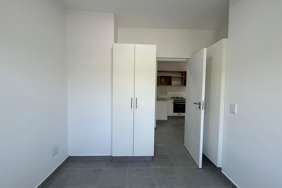 A clean, empty bedroom featuring neutral tiled flooring and a built-in wardrobe, with a view through to the kitchen area.