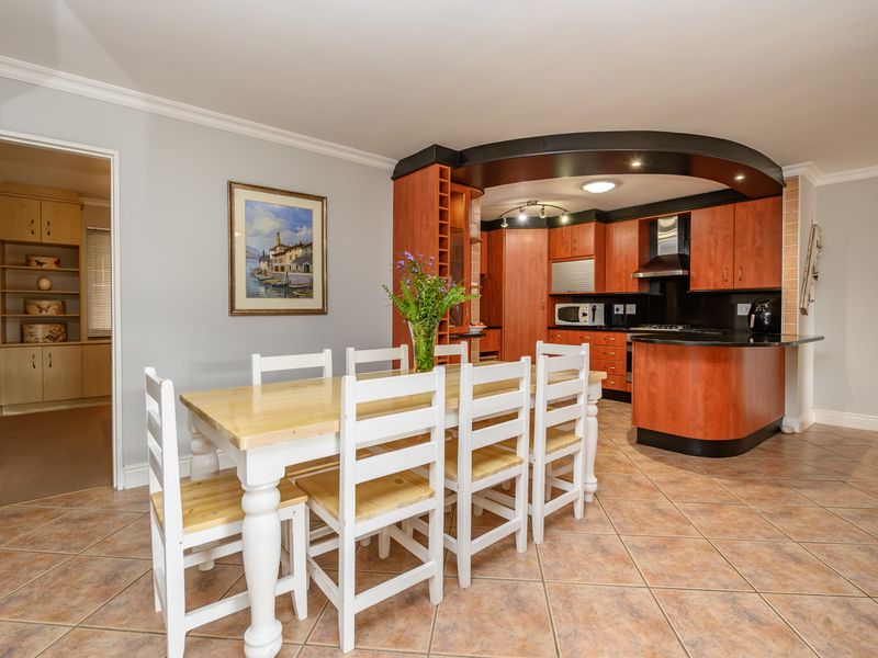 Open-plan kitchen and dining area featuring warm-toned cabinetry, dark countertops, integrated appliances, and tiled flooring, with an adjacent walk-in pantry or scullery space.