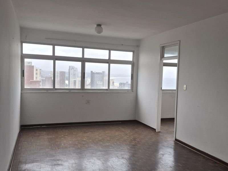 An empty living room featuring original parquet flooring, large windows with city views, and neutral wall finishes, offering a blank canvas for renovation.