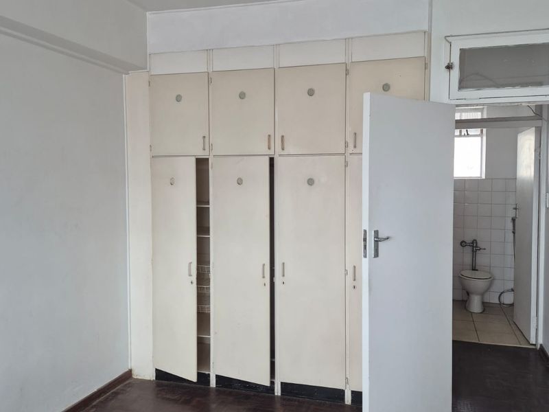 A vacant bedroom featuring original parquet flooring and extensive built-in cupboard storage, with an adjacent bathroom visible through the doorway.