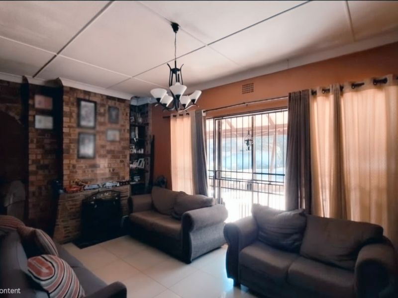 A living room featuring an exposed brick fireplace, tiled flooring, and access to an outdoor area through a sliding glass door with security gate.