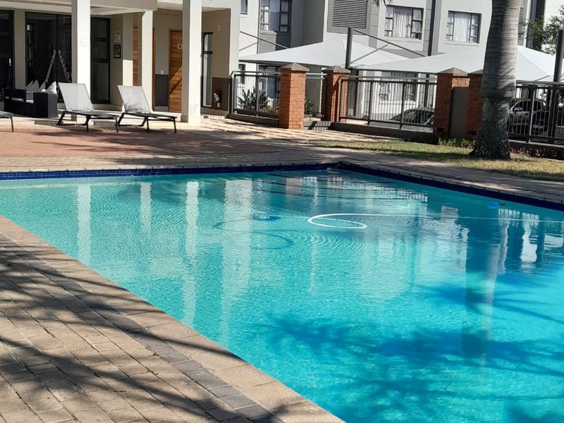 A well-maintained communal swimming pool area featuring paved decking, brick boundary walls, and access to a covered patio space within the apartment complex.