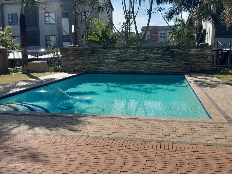 A well-maintained communal swimming pool area featuring a decorative stone feature wall, paved surround, and established landscaping within an apartment complex.