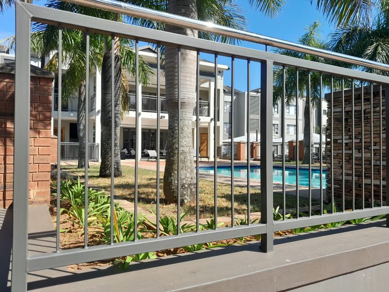 A view of the apartment complex communal swimming pool area, featuring paved decking and secure metal perimeter fencing.