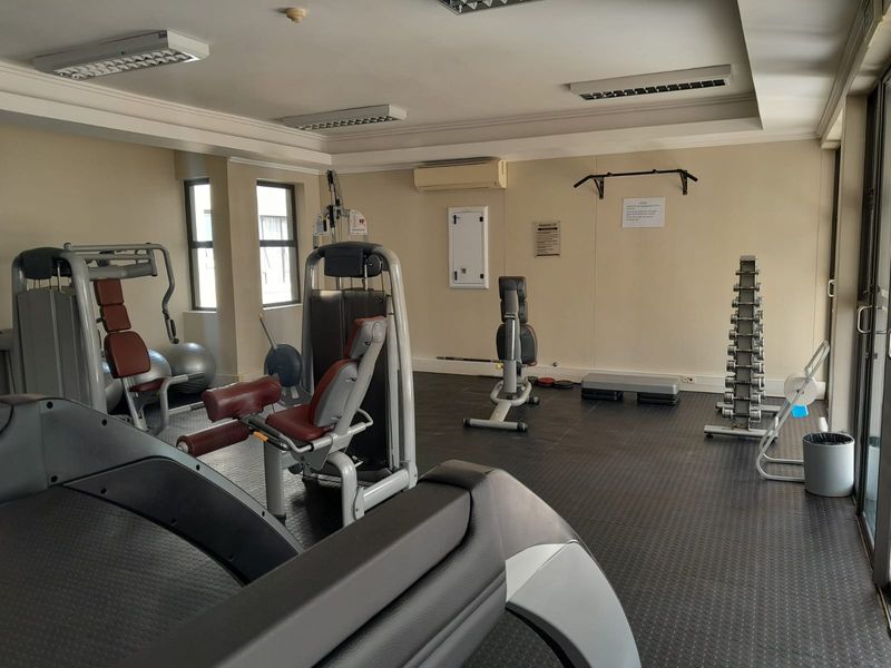 A well-maintained apartment complex gym featuring rubber flooring, wall-mounted air conditioning, and various fixed exercise equipment.