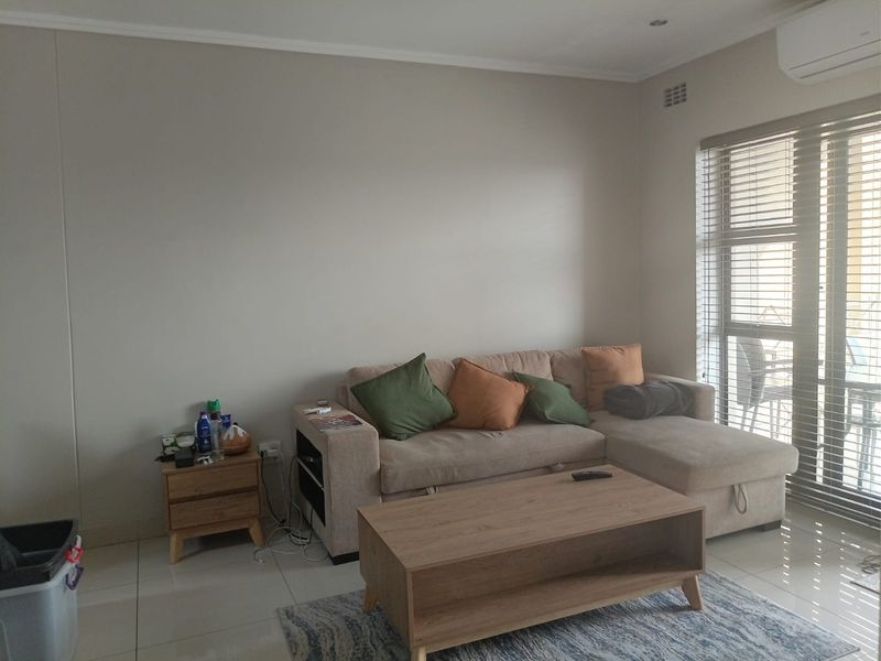 A well-lit living room featuring tiled flooring, an integrated air conditioning unit, and large windows fitted with venetian blinds.