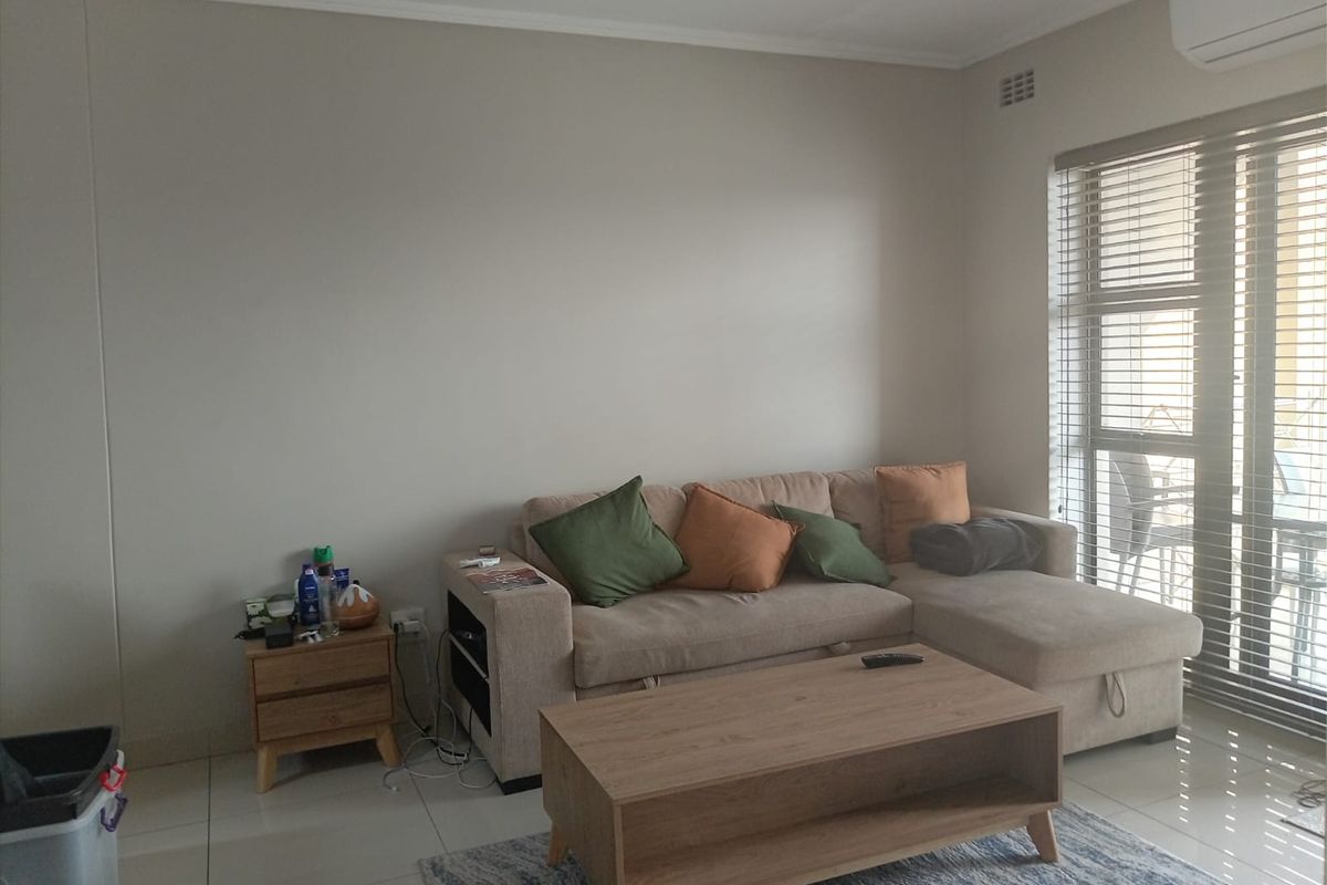 A well-lit living room featuring tiled flooring, an integrated air conditioning unit, and large windows fitted with venetian blinds.