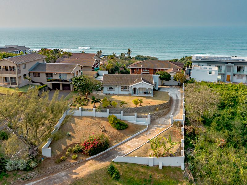 Aerial view of a coastal residential property featuring a tiled roof house, tiered garden, concrete driveway, and perimeter walling with ocean views.