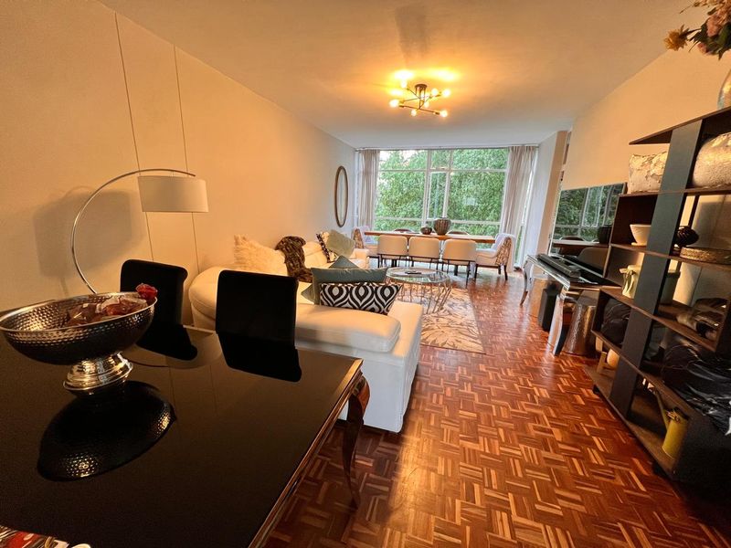 Spacious open-plan living and dining area featuring classic parquet flooring and large windows that provide ample natural light and a pleasant green outlook.