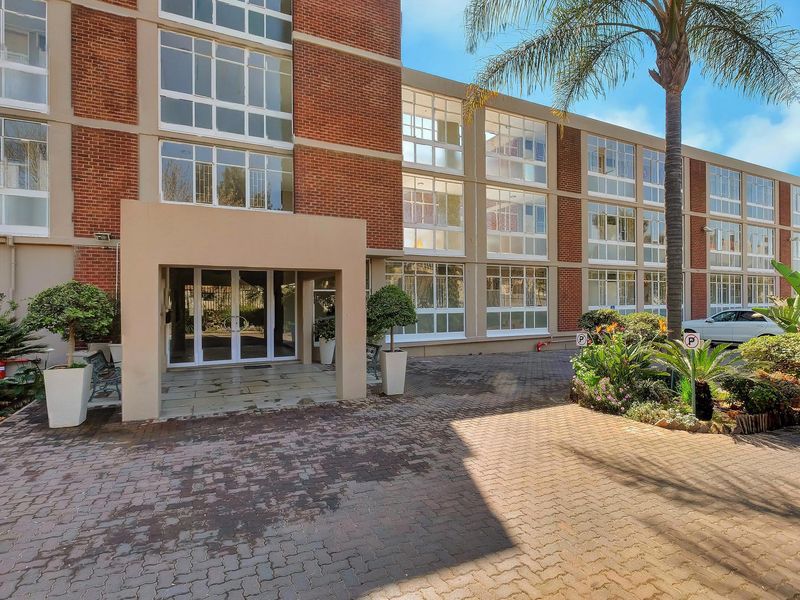 Front exterior view of a multi-story apartment building featuring a brick facade, large windows, and a paved entrance area with landscaped surroundings.