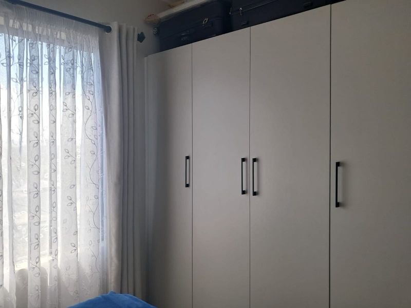 A bedroom featuring a modern, neutral-toned built-in wardrobe with sleek black handles and a window with sheer curtains.