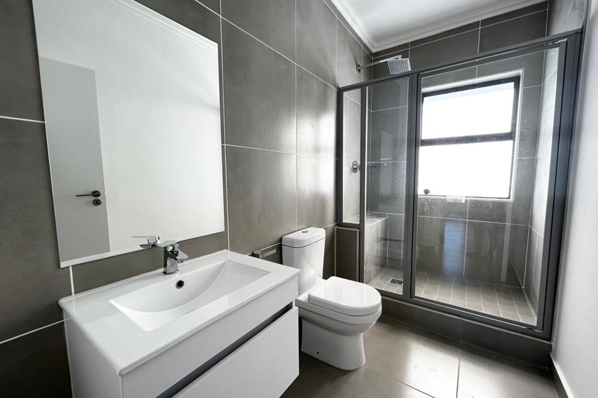 Modern bathroom featuring a sleek vanity, wall-mounted mirror, toilet, and a glass-enclosed shower with contemporary grey wall and floor tiling.