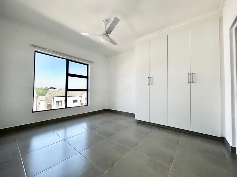 A bright, empty bedroom featuring large format floor tiles, built-in white cupboards, and a ceiling fan with a view of the surrounding estate.