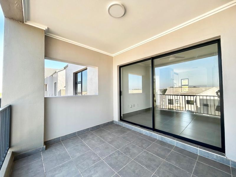A clean, modern covered balcony featuring tiled flooring, a large sliding glass door, and a metal balustrade, offering a bright outdoor space.
