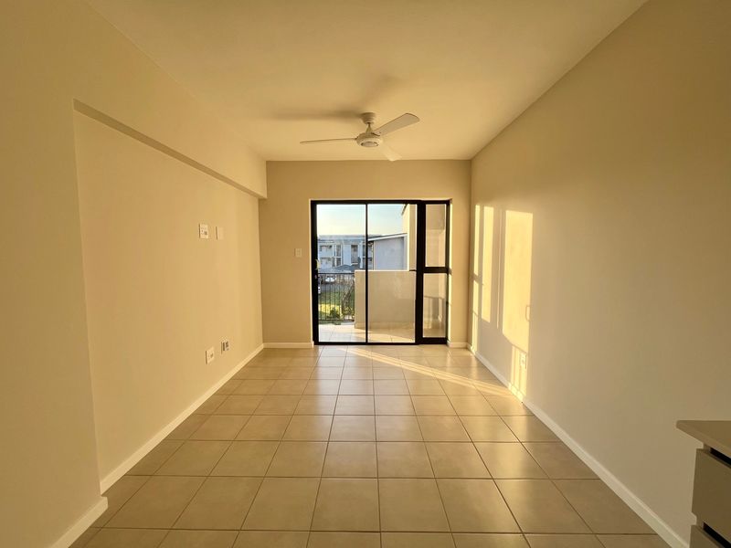 A bright, empty living area featuring neutral tiled flooring, a ceiling fan, and a sliding glass door leading to an outdoor balcony.